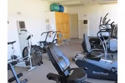 Interior view of a fitness room with various exercise equipment including stationary bikes, an elliptical machine, and exercise balls placed on a shelf against the wall.