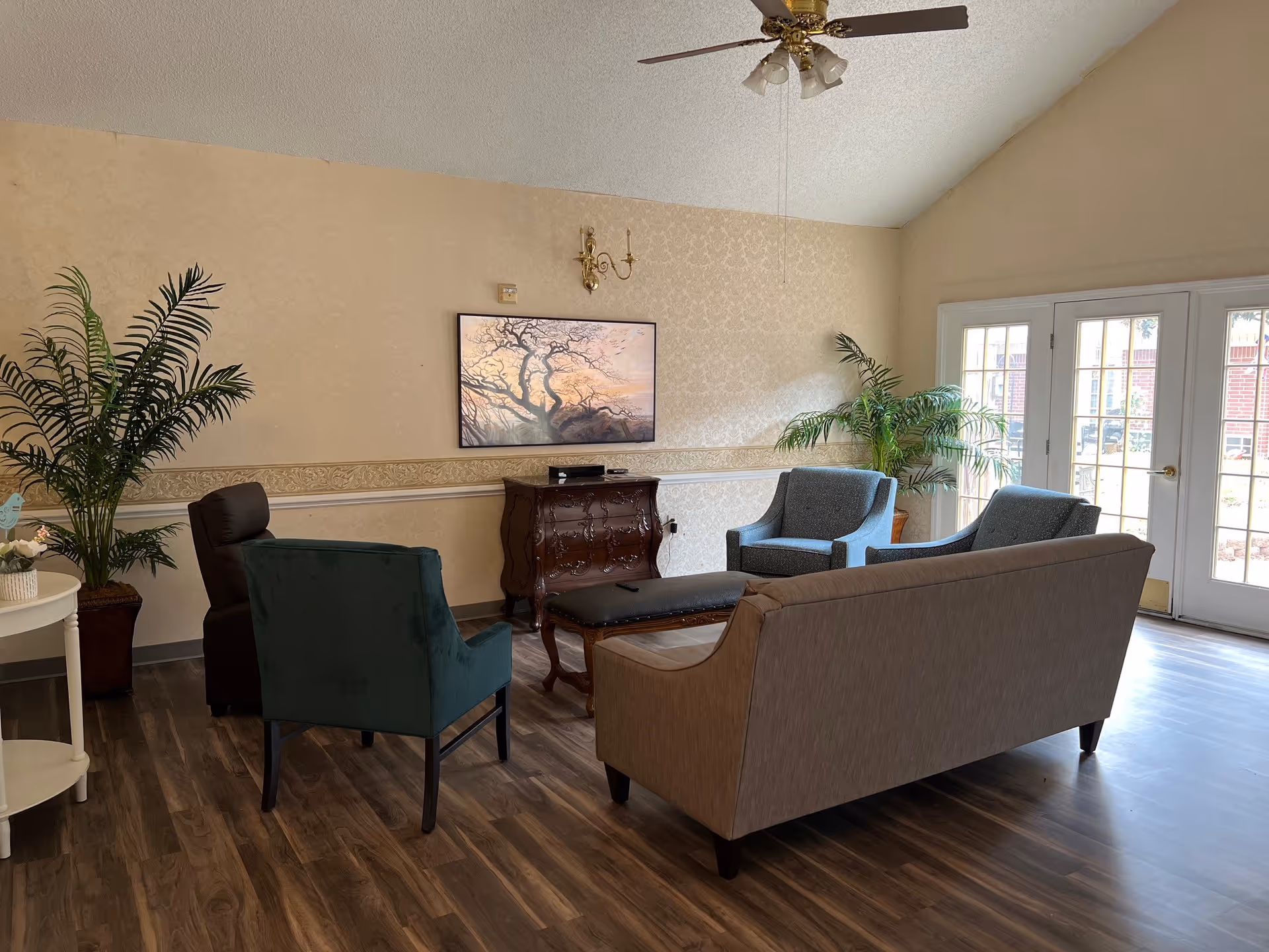 A cozy living room area with a beige couch, two blue armchairs, a dark green armchair, and a dark brown leather chair arranged around a wooden coffee table. The room has wood flooring, cream-colored wallpaper with a decorative border, two large potted plants, a ceiling fan with lights, and a wall-mounted flat screen TV above a wooden cabinet. French doors with glass panes lead outside, allowing natural light to fill the room.