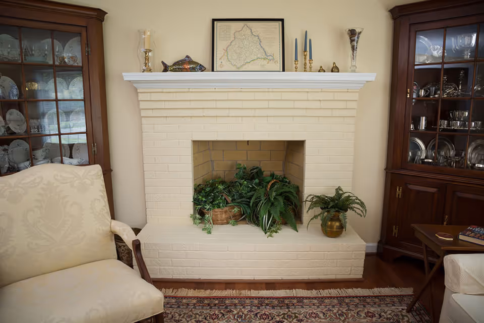 A cozy living room area featuring a white brick fireplace filled with green plants. On the mantel above the fireplace are decorative items including a framed map, candlesticks, and a fish sculpture. Flanking the fireplace are two wooden display cabinets filled with dishes and silverware. A beige upholstered armchair is positioned to the left, and a small wooden side table with books is on the right. The floor has a patterned rug.