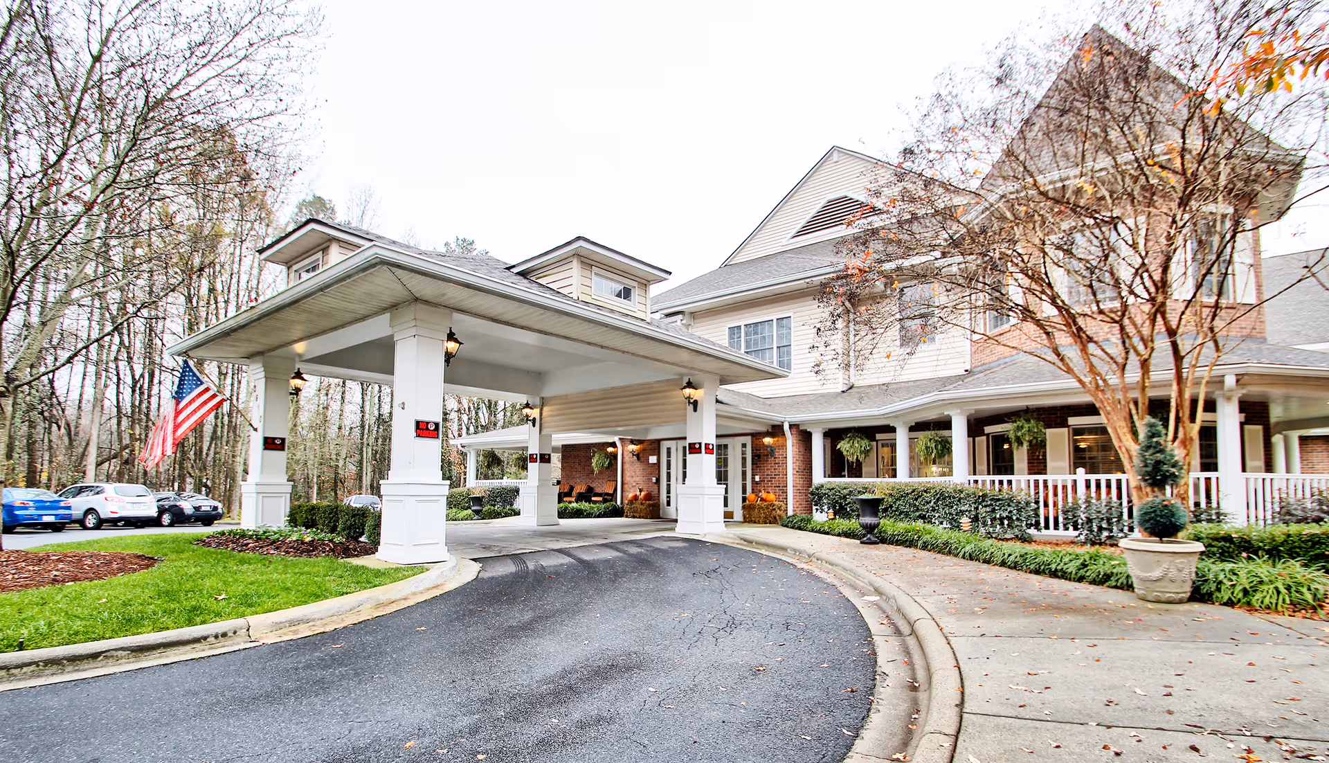 Exterior view of Charter Senior Living of Charlotte showing the entrance with a covered drop-off area, surrounded by trees and parked cars. The building has a porch with white columns and hanging plants, and an American flag is displayed near the entrance.