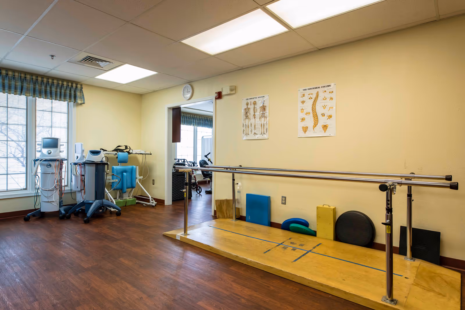 Physical therapy room with parallel bars on a wooden platform, exercise equipment, anatomical posters of the skeletal system and vertebral column on the wall, and a large window with blue checkered curtains.