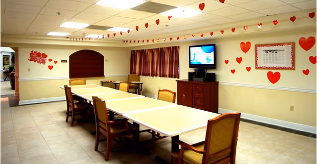 A long rectangular table with several chairs around it in a room decorated with red heart-shaped cutouts on the walls and hanging from the ceiling. There is a flat-screen TV mounted on the wall above a wooden cabinet, and a window with striped curtains. The room has tiled floors and a drop ceiling with fluorescent lighting.