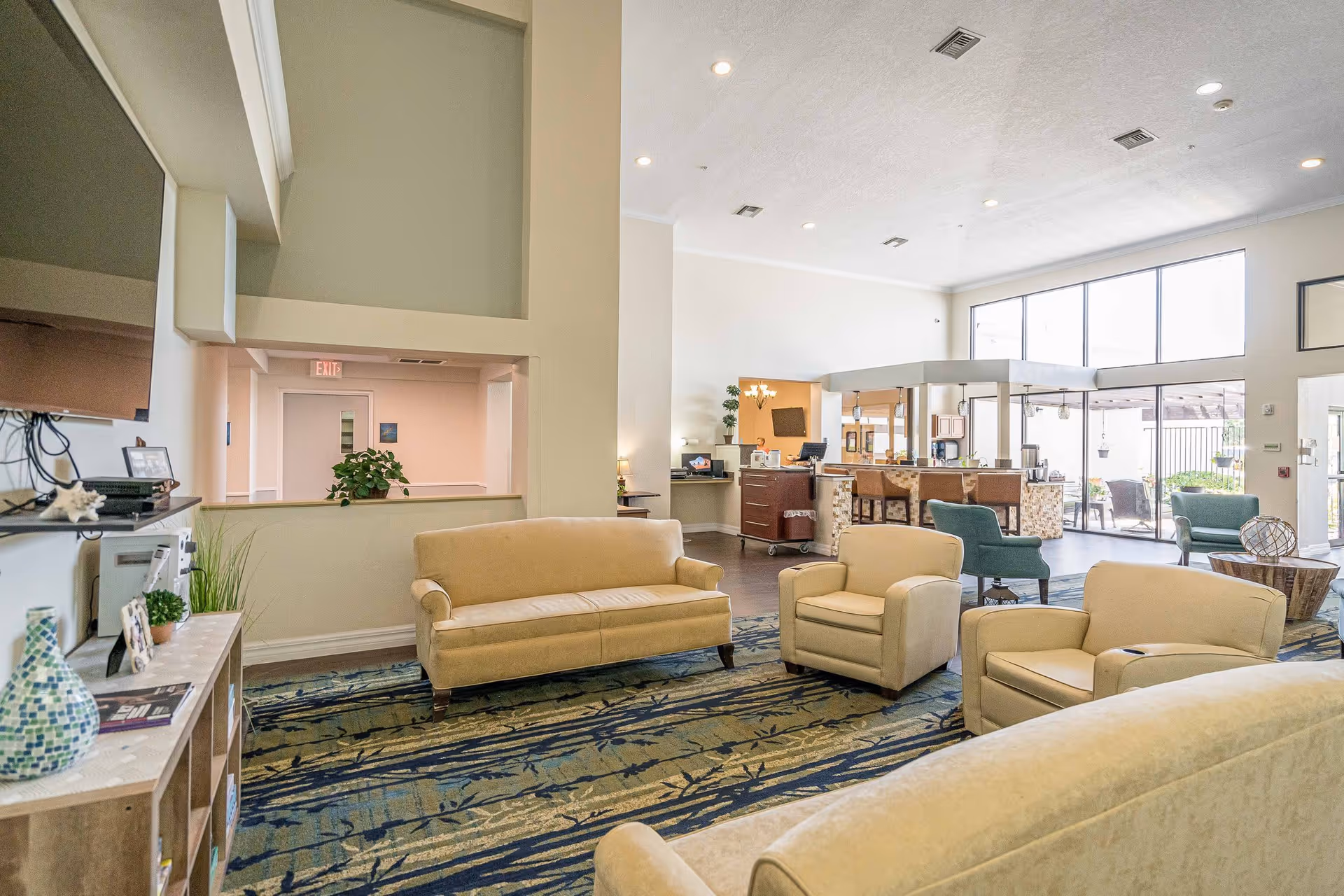 A spacious and bright common area in an assisted living facility featuring beige sofas and armchairs arranged on a patterned carpet. The room has high ceilings with recessed lighting, large windows allowing natural light, and a reception or service desk area in the background with bar stools. There are plants and decorative items on shelves and tables, creating a welcoming atmosphere.