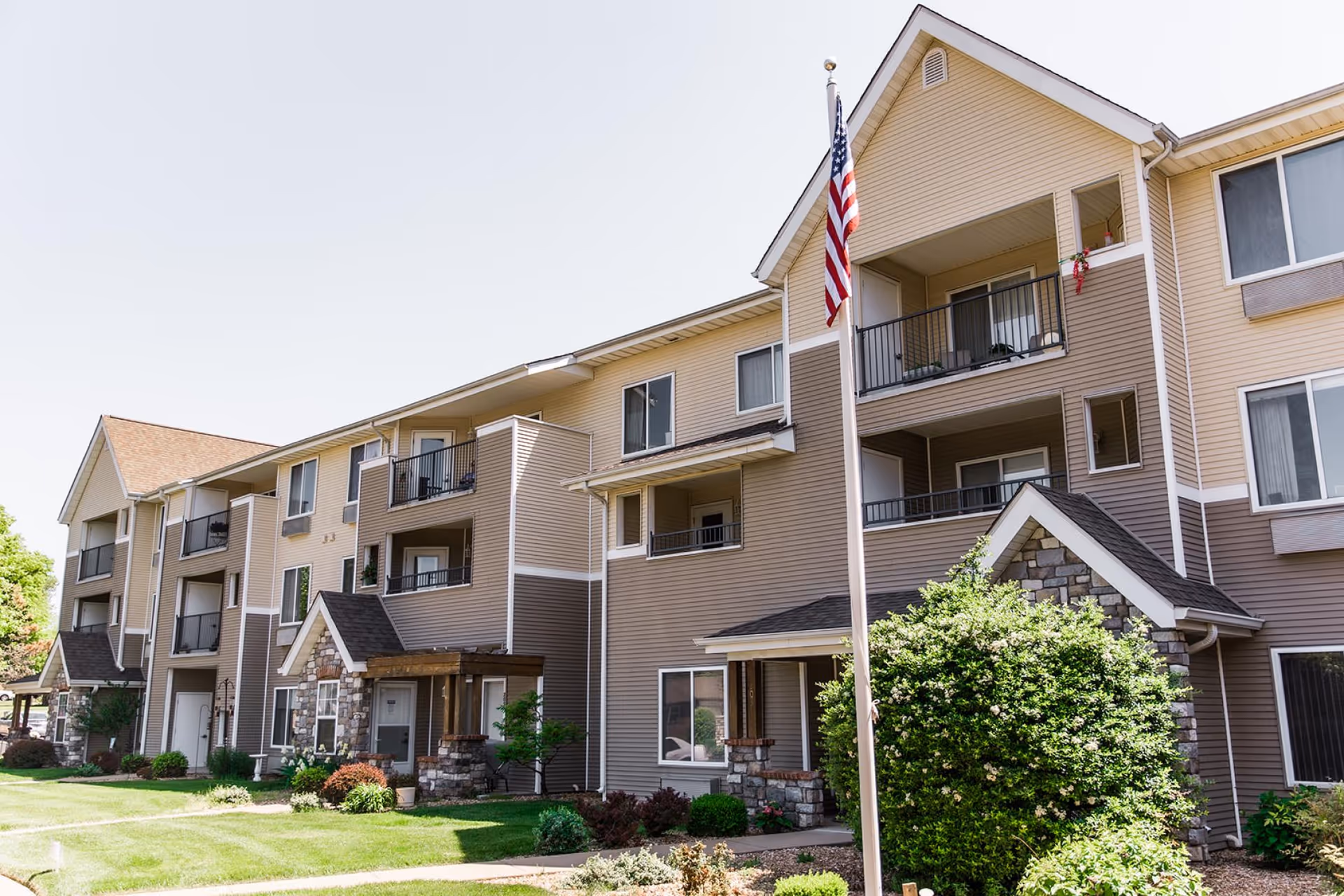 Front exterior of a multi-story beige senior living apartment building with balconies, landscaping, and an American flag.