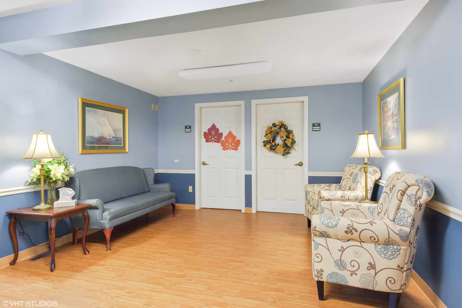 A cozy sitting area in a senior living facility with light blue walls and wooden flooring. The room features a blue upholstered sofa on the left, a wooden side table with a lamp and flowers, and two patterned armchairs with a lamp between them on the right. Two white doors decorated with autumn-themed wreaths and leaves are visible in the background. Framed paintings hang on the walls above the seating.