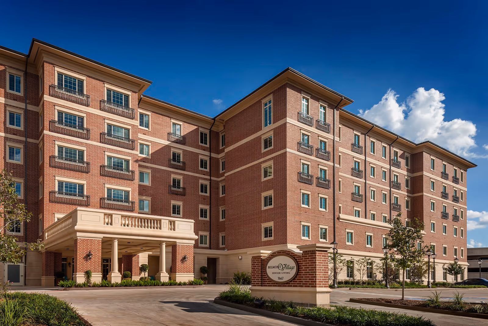Exterior view of Belmont Village Senior Living Hunters Creek, a multi-story brick building with numerous windows and a covered entrance. The building is surrounded by landscaped greenery and a clear blue sky with some clouds.