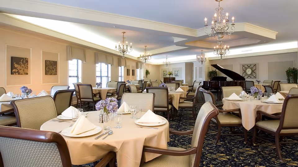 Elegant dining room with round tables covered in beige tablecloths, each set with folded white napkins, glassware, and silverware. The room features upholstered chairs, chandeliers hanging from the ceiling, large windows with beige valances, decorative wall art, and a grand piano in the background.