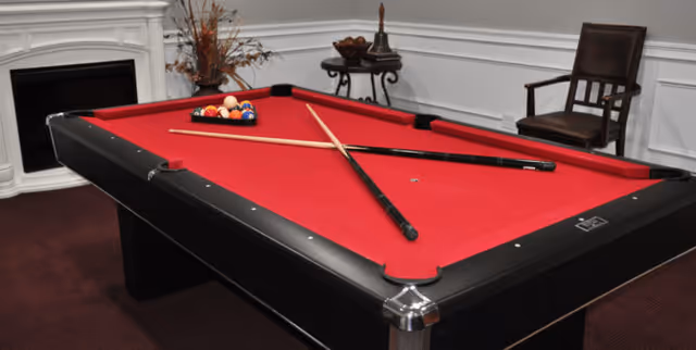 A red felt pool table with two crossed pool cues and a rack of billiard balls set up on the table. The room has white paneled walls, a fireplace on the left, a small round table with decorative items in the corner, and a wooden chair on the right.