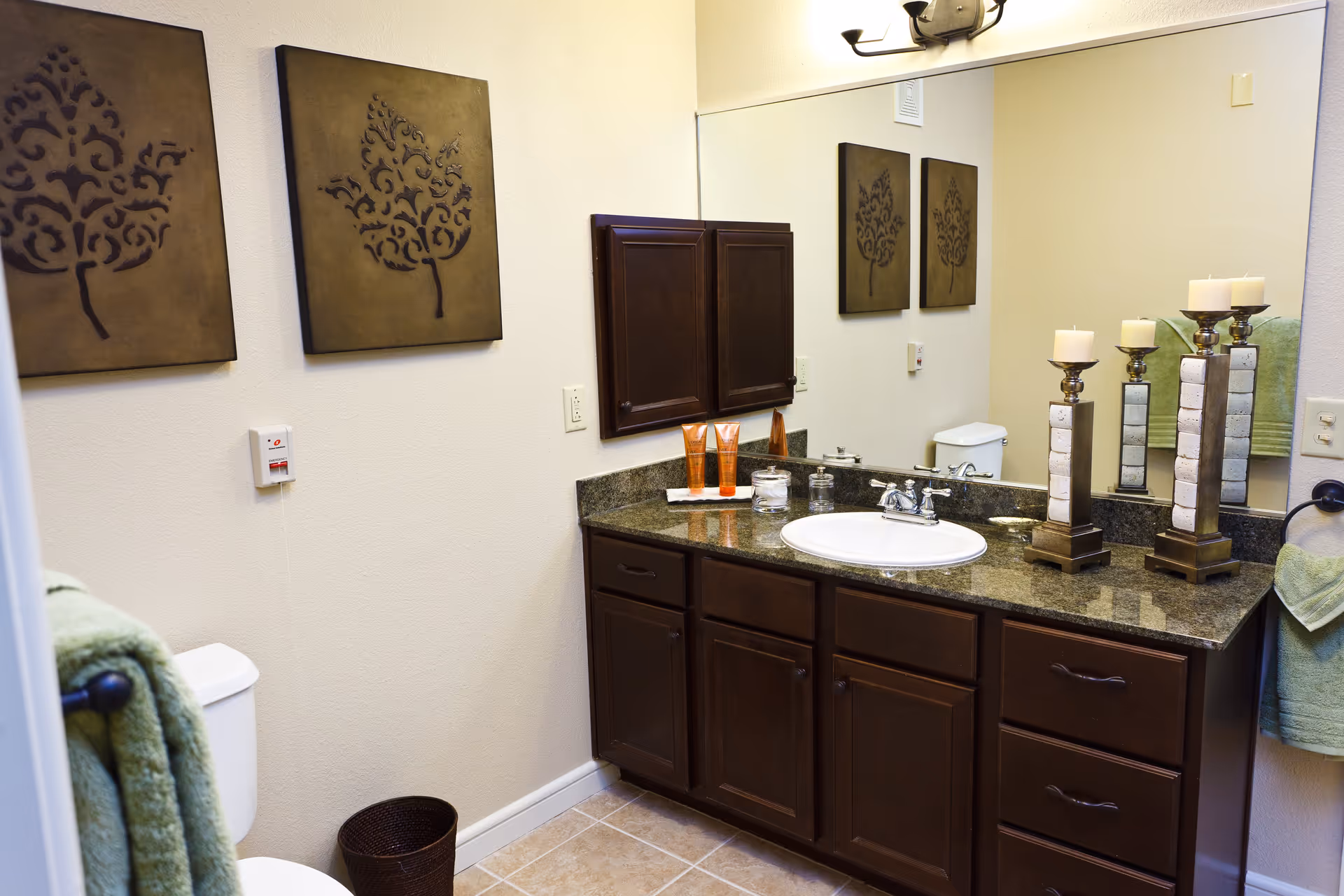 A bathroom with a granite countertop vanity featuring a white sink and dark wooden cabinets. Above the vanity is a large mirror reflecting two decorative candle holders with white candles. On the wall above the toilet are two framed artworks with tree designs. The bathroom has beige tiled flooring and light-colored walls, with green towels hanging on towel racks.