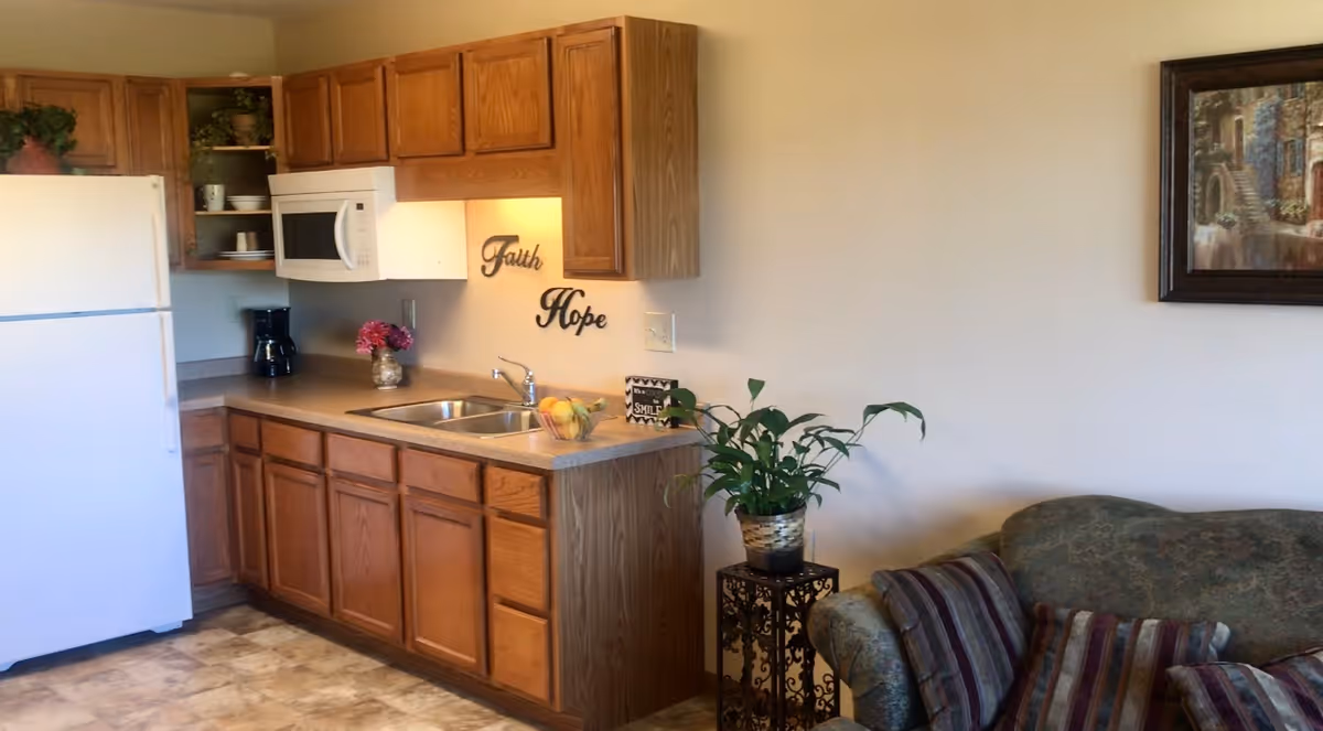 A small kitchen area with wooden cabinets, a white refrigerator, a microwave, a coffee maker, and a sink. On the wall above the sink are decorative words 'Faith' and 'Hope'. To the right of the kitchen is a green patterned couch with striped pillows and a small plant on a decorative stand. A framed painting hangs on the wall above the couch.