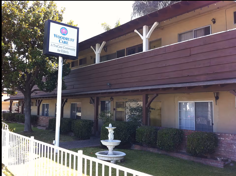 Exterior view of Woodruff Care, a two-story elderly care community building with beige walls and brown wooden balcony railing. There is a white fence in the foreground, a small white fountain on the grass, and a signpost with the Woodruff Care logo and text reading 'A TruCare Community for Elderly'.