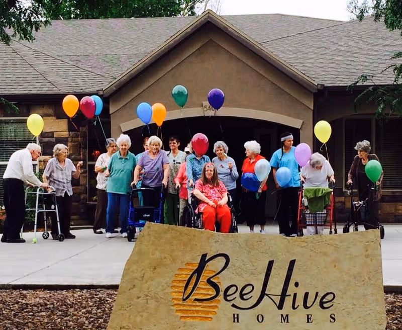 A group of elderly residents holding colorful balloons stand in front of the Beehive Homes building with a large stone sign in the foreground.