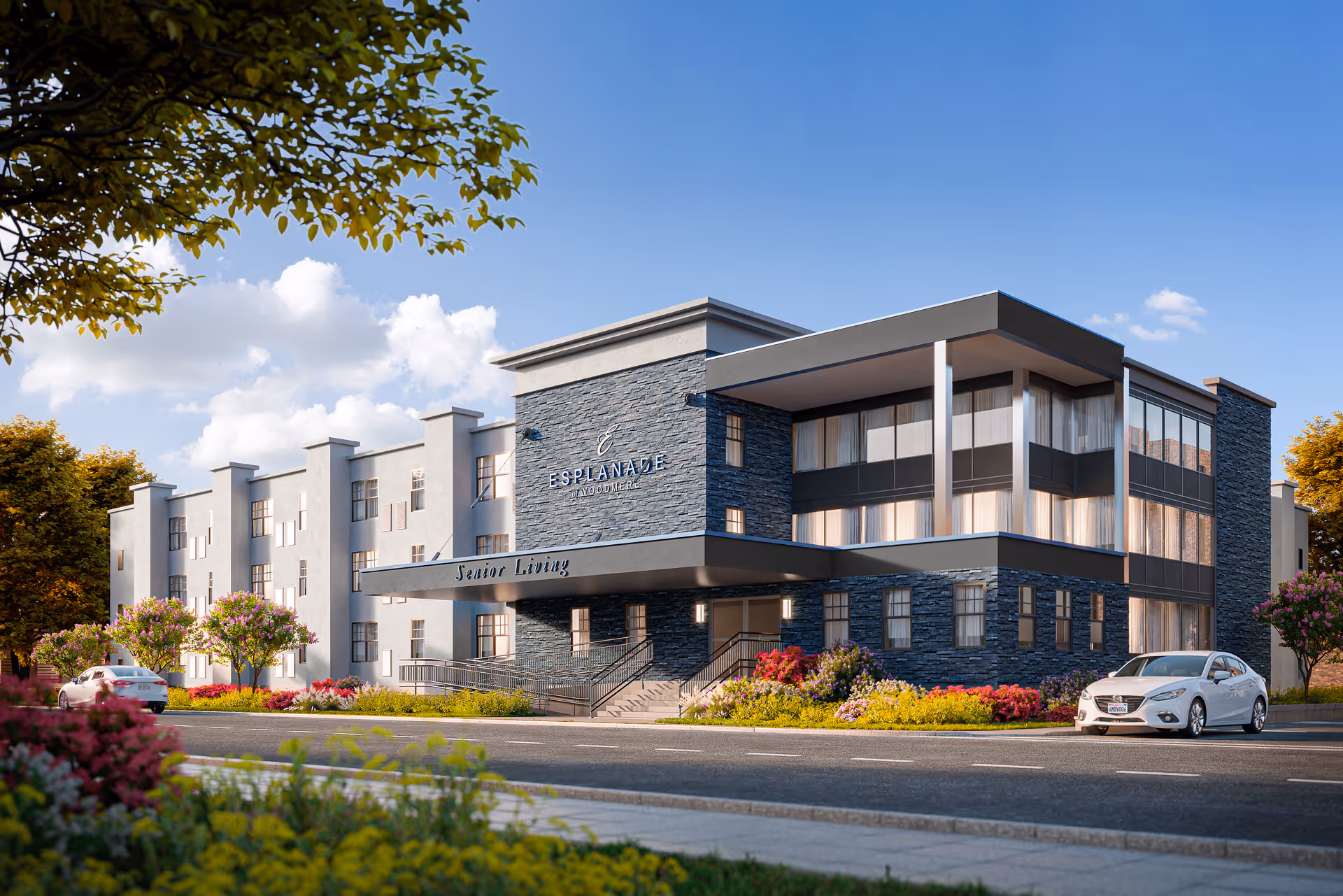 Exterior view of Esplanade of Woodmere senior living facility building with modern architecture, stone and white walls, large windows, landscaping with flowers and trees, and a white car parked in front under a clear blue sky.