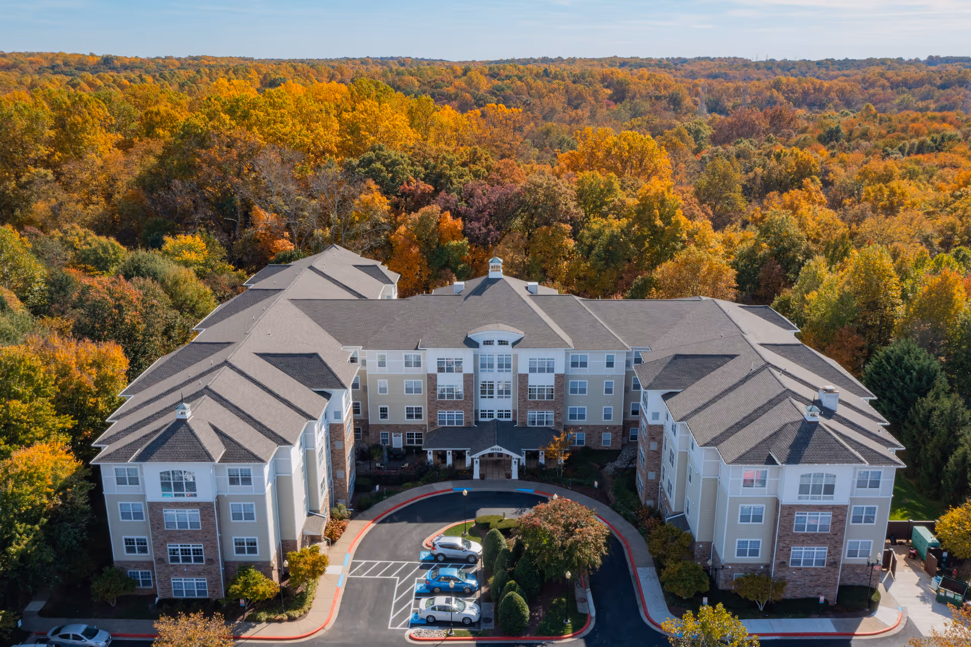 Aerial view of a large senior living facility building surrounded by dense trees with autumn foliage in vibrant shades of orange, yellow, and green. The building has multiple stories with a central entrance and a circular driveway with parked cars. The facility is named Willow Manor At Cloppers Mill (Seniors 62+).