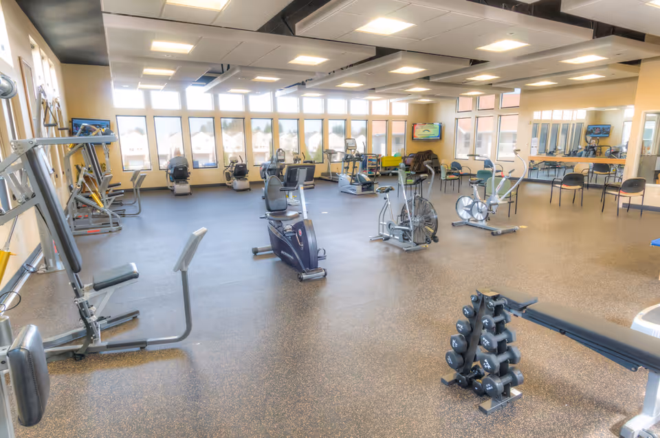 A spacious fitness room with various exercise equipment including stationary bikes, weight machines, dumbbells, and elliptical machines. The room has large windows letting in natural light, a television mounted on the wall, and several chairs arranged along the walls.