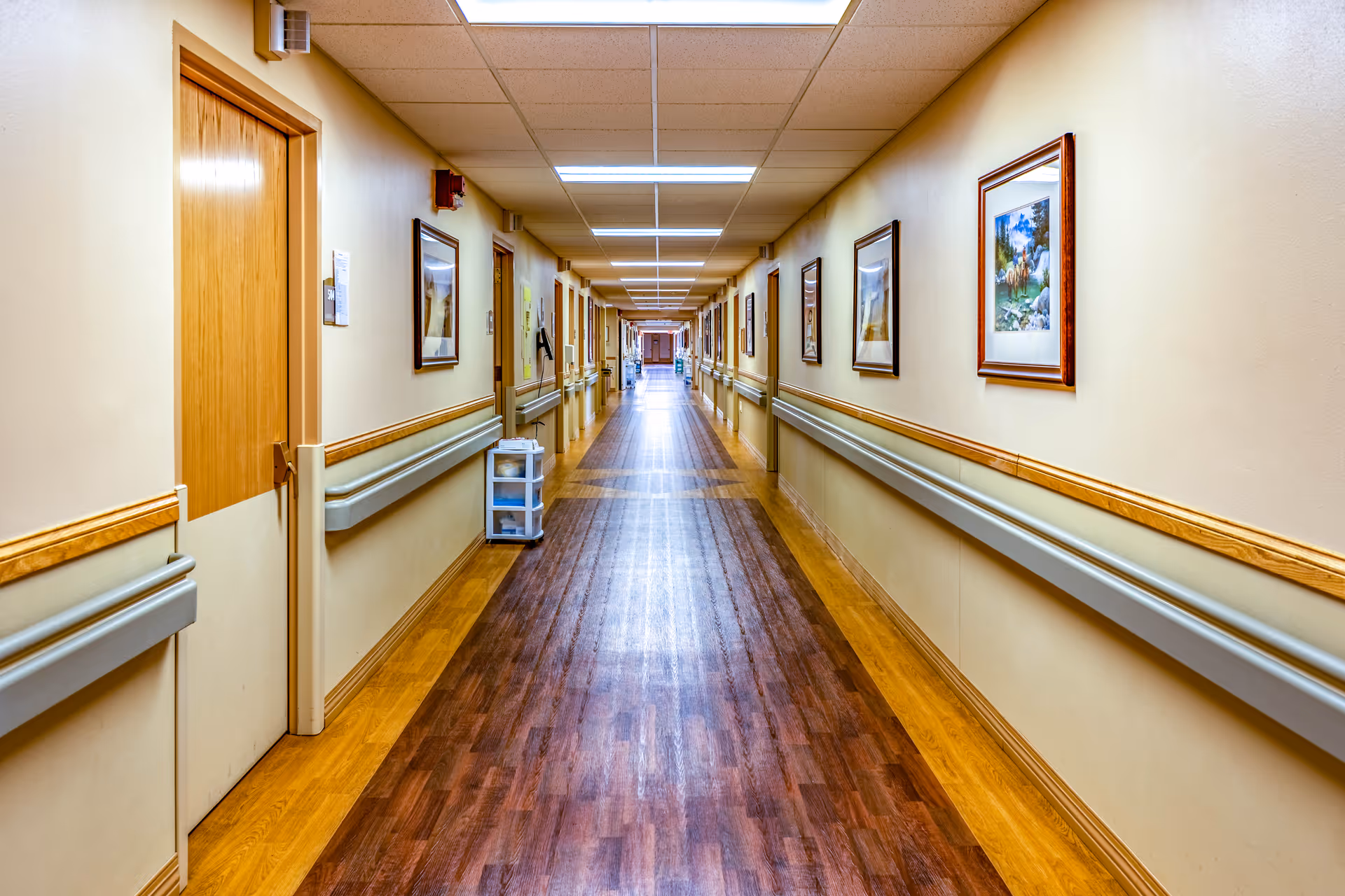 Long, well-lit hallway in a senior living facility with wood floors, handrails, framed artwork, and doors along both sides.