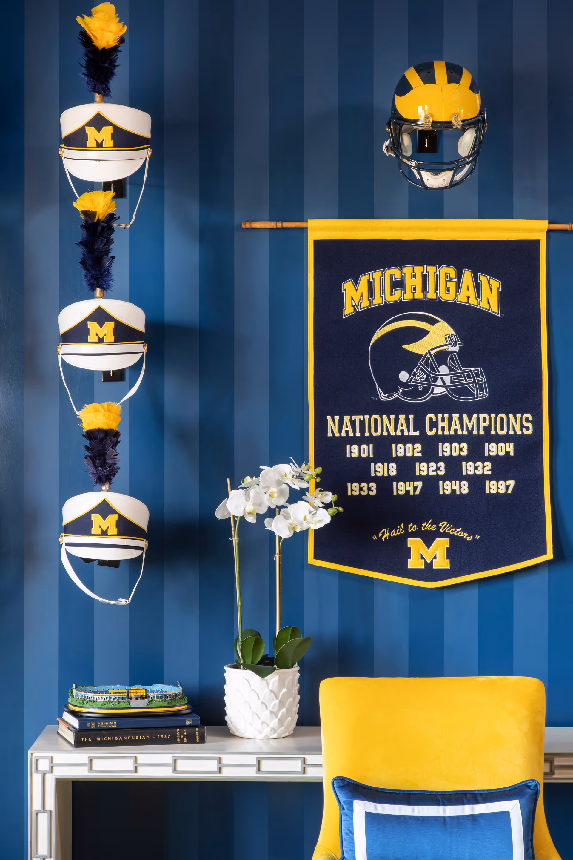 A blue striped wall decorated with three white and blue marching band hats with yellow and blue plumes on the left, a yellow and blue football helmet mounted on the right, and a blue and yellow banner listing Michigan National Champions years. Below is a white table with books, a model stadium, and a white orchid plant. In front is a yellow chair with a blue and white pillow.