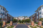 Front exterior of a multi-story brick and siding senior living building with a circular driveway, landscaped courtyard, and lampposts under a clear blue sky.