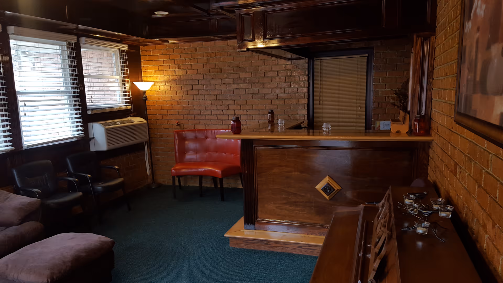 Interior lounge with exposed brick walls, a wooden bar, red upholstered bench, armchairs and windows with blinds.