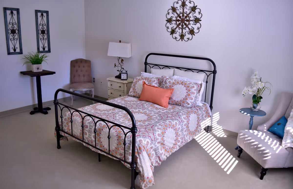 A neatly made bed with a black metal frame and patterned bedspread featuring orange and white designs. There is a coral-colored rectangular pillow in the center. To the left of the bed is a small white nightstand with a lamp and a clock. A tufted beige chair is positioned in the corner near a small table with a potted plant. On the right side of the bed, there is a small round blue table with a white orchid plant and a beige armchair with a blue cushion. Decorative wall art hangs above the bed and on the left wall. Sunlight streams through blinds, casting shadows on the floor and chair.