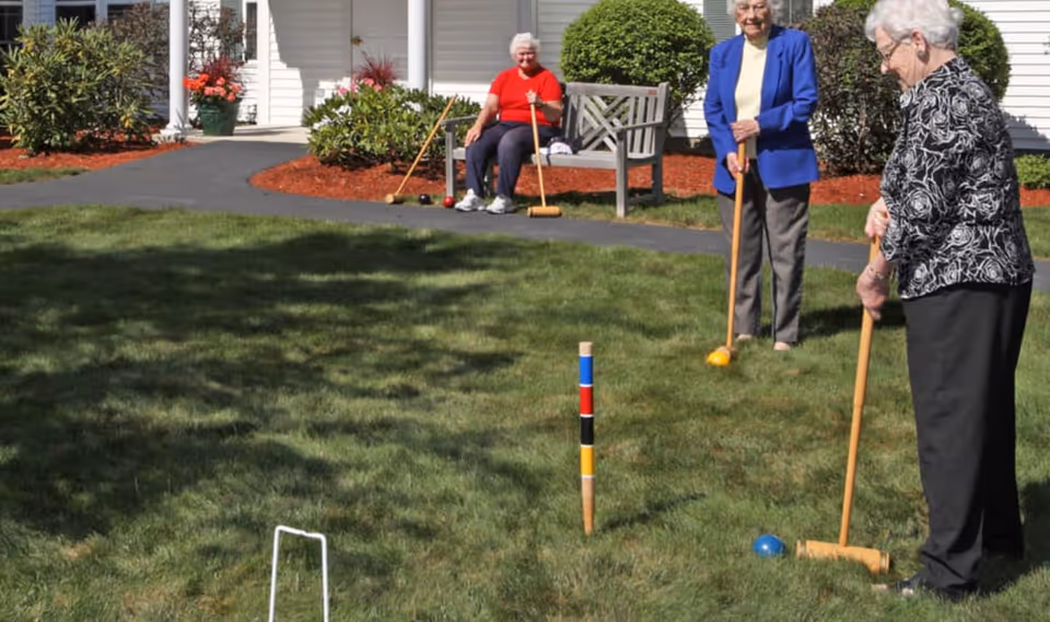 Three elderly women playing croquet on a grassy lawn outside a white building with shrubs and a bench. One woman is sitting on the bench wearing a red shirt, while the other two women stand holding croquet mallets, one in a blue jacket and the other in a black and white patterned top.