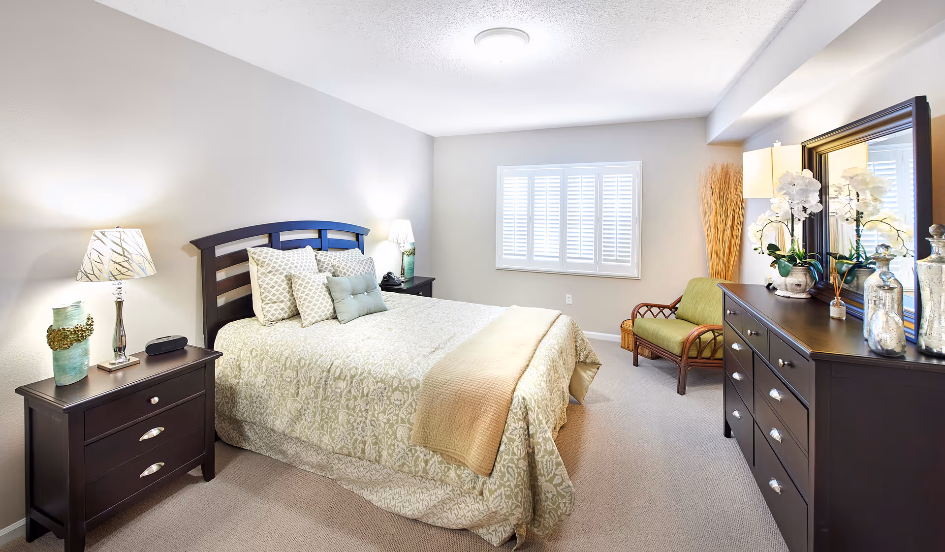 Bright, neatly staged bedroom with a bed, two nightstands, a dresser with mirror, and a small armchair.