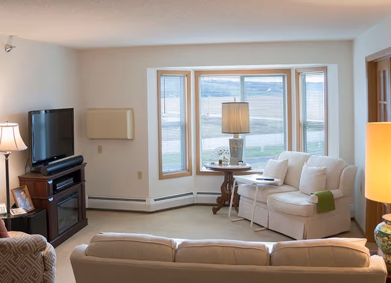 Bright living room with sofas, a TV on a stand, a bay window with blinds, and a side table with a lamp.