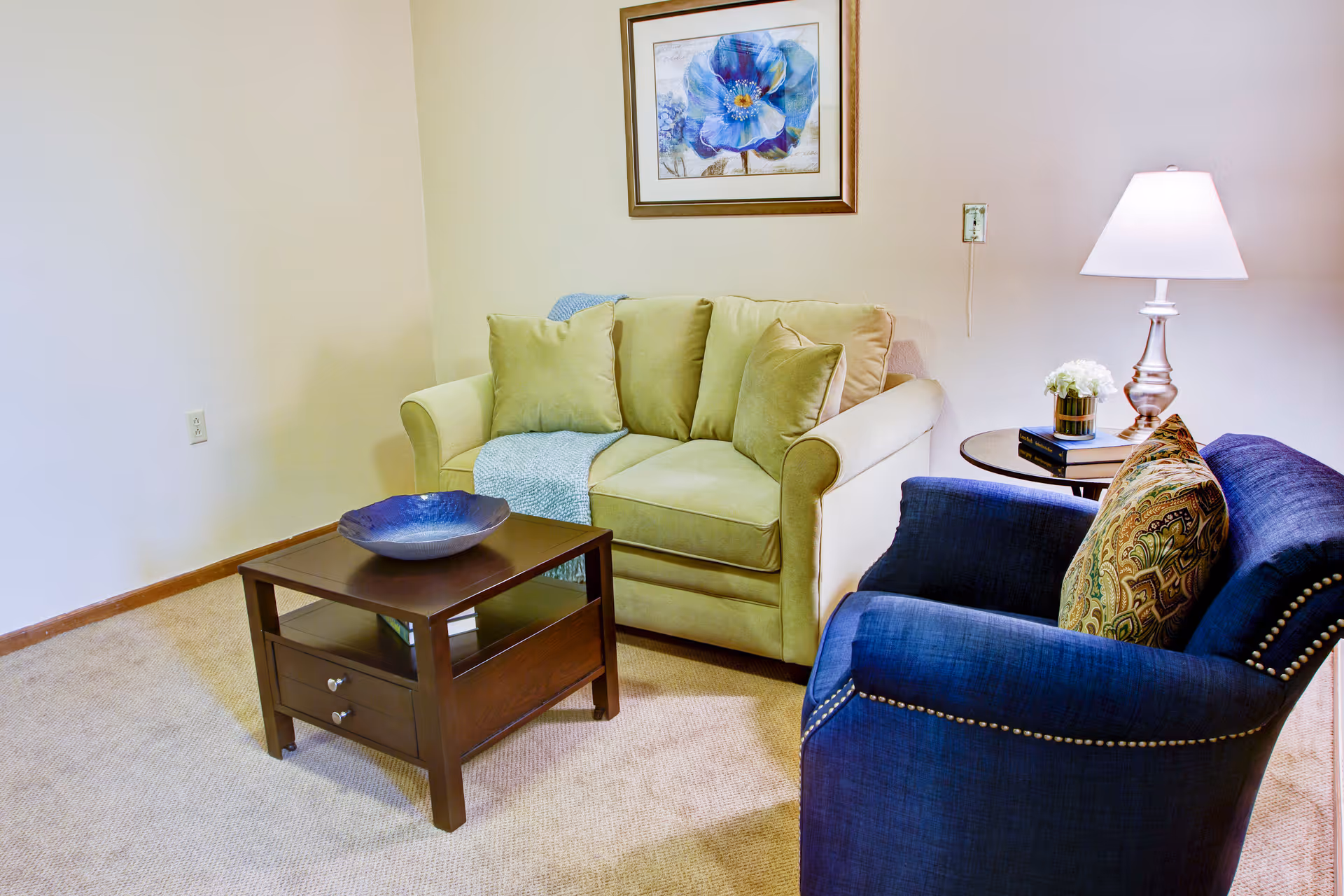 Small living room with a green loveseat, blue armchair, wooden coffee table, side table with a lamp, and a framed floral print on the wall.
