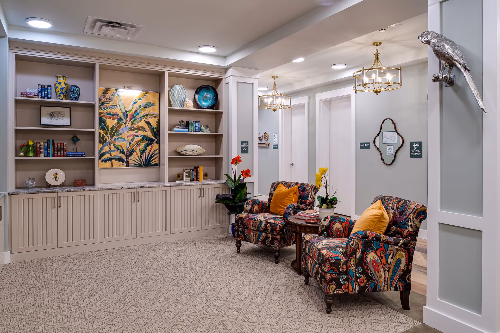 A cozy sitting area in a senior living facility featuring two colorful patterned armchairs with orange and yellow pillows, a small round wooden table with books and a potted orchid, built-in shelves with decorative items and books, a large painting of tropical leaves, and a silver parrot wall sculpture. The space is well-lit with ceiling lights and gold chandeliers, and there are doors labeled 'Laundry' and other signs in the background.