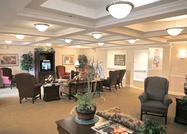 A spacious living room area in a senior living facility with multiple upholstered armchairs and sofas arranged around coffee tables. The room features beige walls, carpeted floors, framed artwork, potted plants, and ceiling light fixtures. A television is visible in a dark wooden cabinet against the far wall.