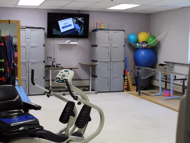 Physical therapy/exercise room with a recumbent bike, storage cabinets, large stability balls, parallel bars and a wall-mounted TV.