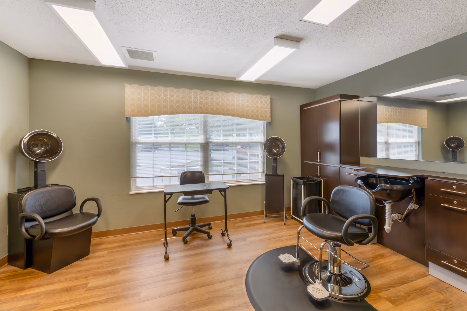 A hair salon room with wooden flooring, two black salon chairs, two hair drying stations, a small black desk with a chair in front of a window with blinds and a beige valance, and dark brown cabinetry with a black sink and large mirror.