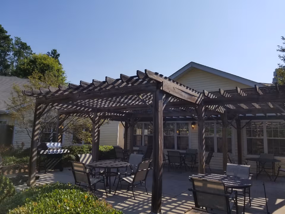 Outdoor patio area with wooden pergolas casting shadows on several tables and chairs. There is a barbecue grill on the left side and a yellow building with windows in the background. Green bushes and trees surround the patio under a clear blue sky.