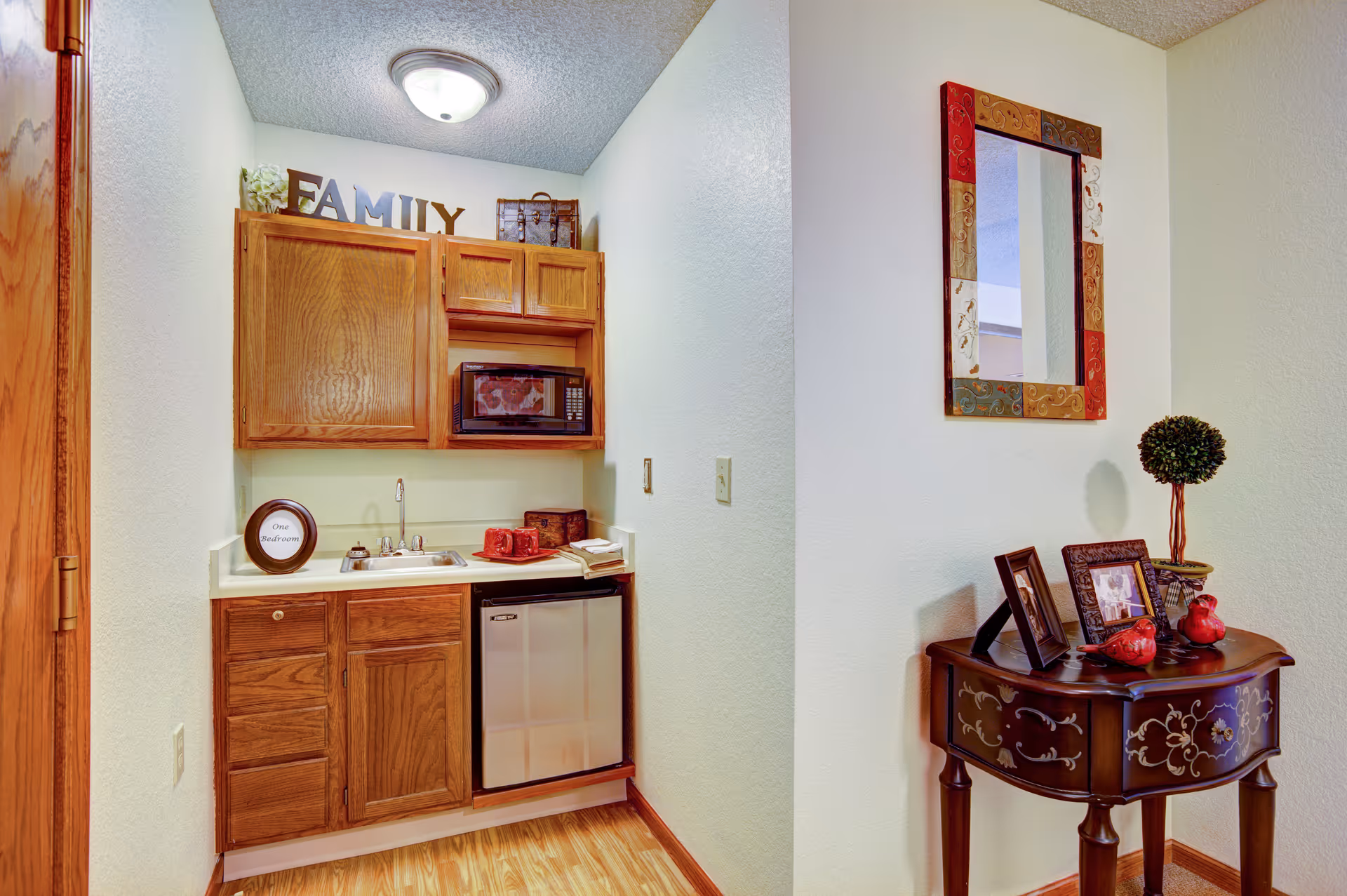 Small kitchenette with wooden cabinets, a sink and mini-fridge beside a decorative table with framed photos and a mirror.