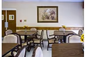 Small dining room with several tables and upholstered chairs, a framed floral painting on the wall, and vases of yellow flowers on the tables.