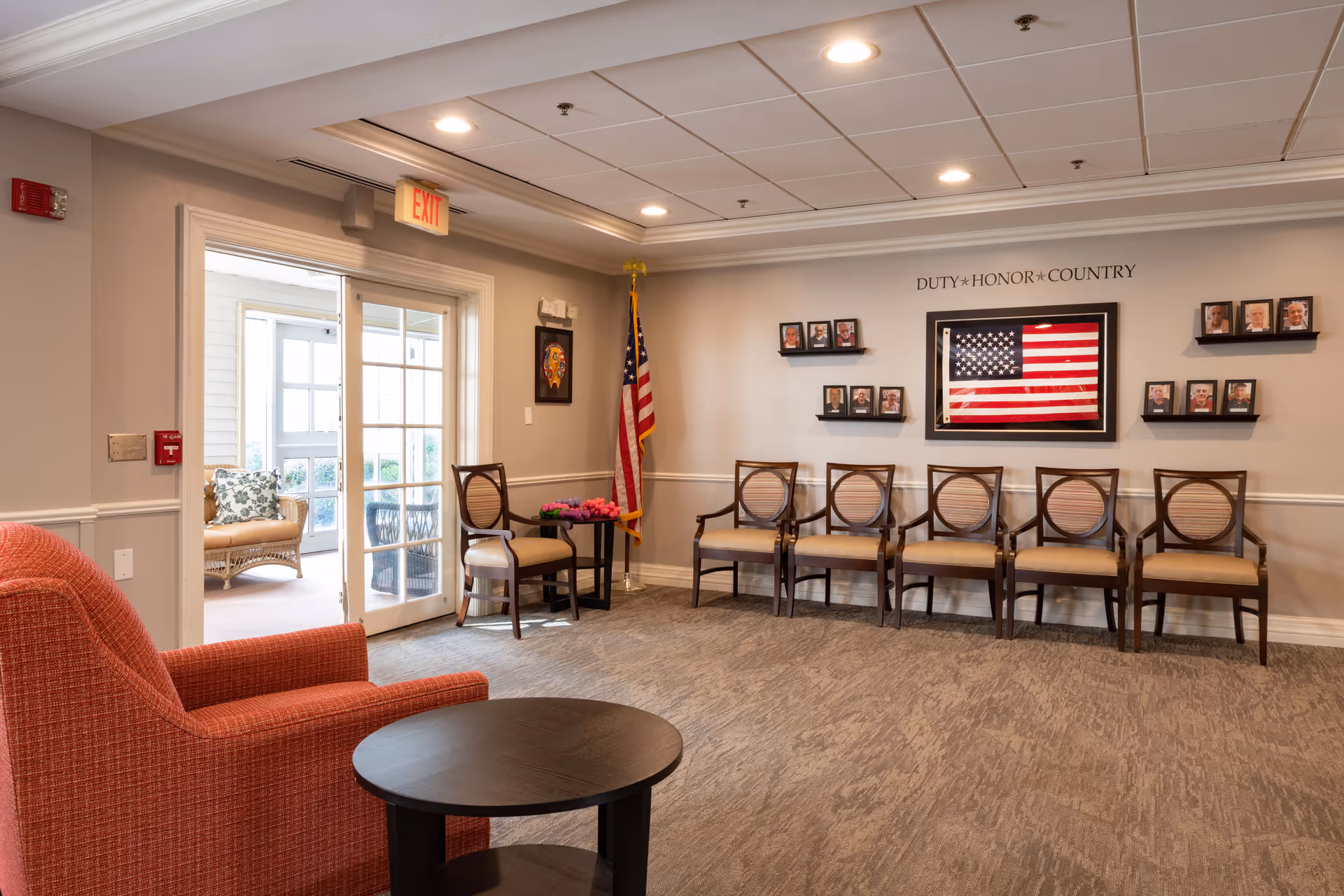 A waiting area in a senior living facility with a row of six wooden chairs with beige cushions against a wall. Above the chairs is a framed American flag and several small framed photos on shelves. The wall has the words 'DUTY * HONOR * COUNTRY' displayed. To the left, there is an American flag on a stand and a small table with flowers. An orange upholstered armchair and a round black table are in the foreground. Double glass doors lead to a sunlit room with wicker furniture. The ceiling has recessed lighting and an exit sign is above the doors.