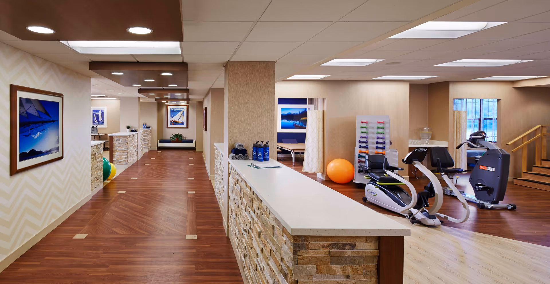 Interior view of a senior living facility fitness and therapy area with exercise equipment including recumbent bikes, a rack of colorful dumbbells, an orange exercise ball, and therapy tables behind privacy curtains. The space features wood flooring, a stone and white countertop reception desk, framed nautical-themed artwork on the walls, and recessed ceiling lights.