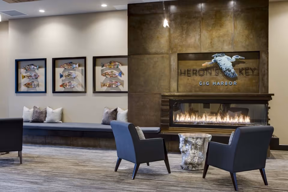 A modern lounge area with two dark armchairs facing a rectangular gas fireplace set in a bronze-toned wall. Above the fireplace is a decorative heron sculpture and the text 'HERON'S KEY GIG HARBOR'. To the left, there is a built-in bench with cushions and three framed artworks featuring colorful fish. The floor is carpeted in neutral tones and the ceiling has recessed lighting.