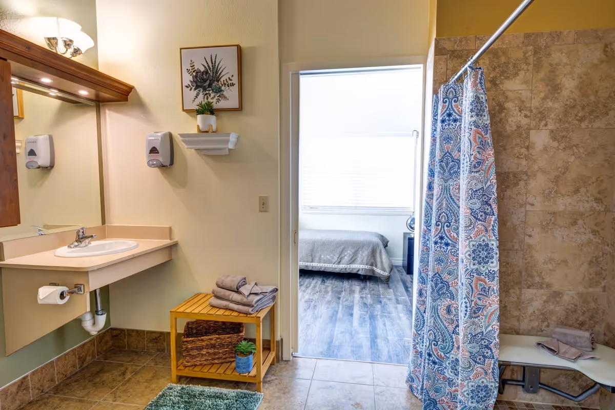 A bathroom with a sink and mirror on the left, a small wooden shelf holding folded towels and a plant, and a shower area with a colorful patterned curtain on the right. Through an open doorway, a bedroom with a bed and a window with blinds is visible.