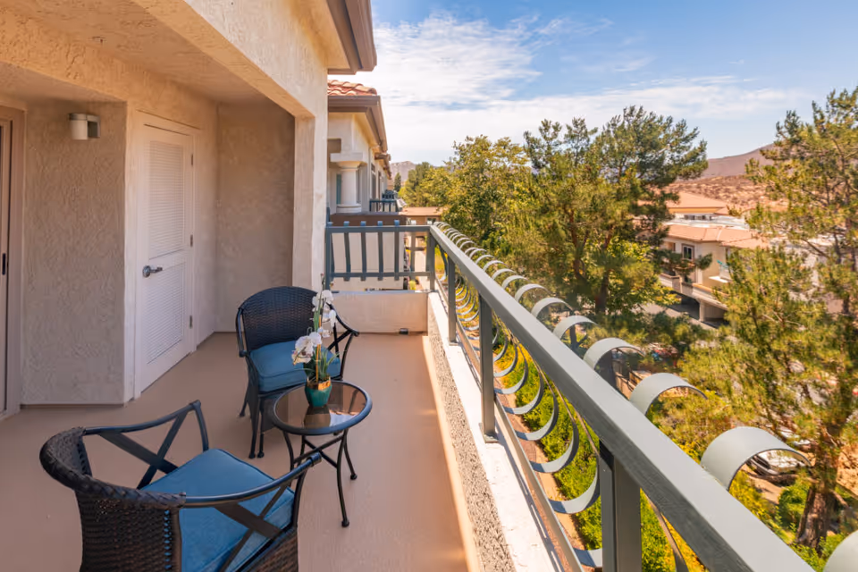 Balcony area with two black wicker chairs with blue cushions and a small round glass table with a potted orchid, overlooking trees and neighboring buildings under a partly cloudy sky.