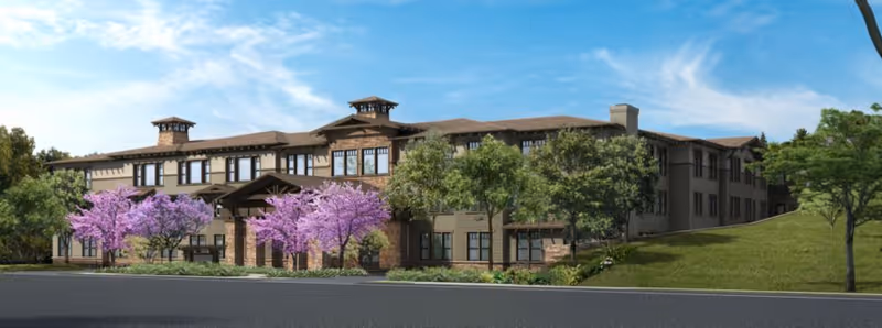 Front exterior of a multi-story senior living building with a covered entrance, landscaped grounds and flowering trees under a blue sky.