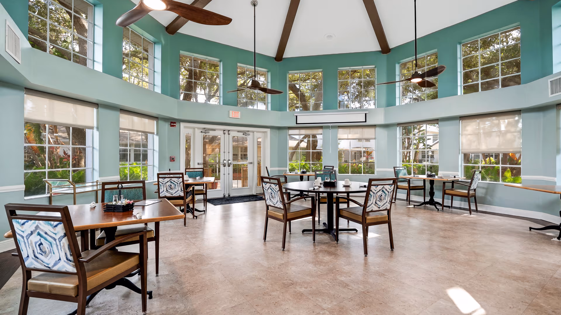 Spacious bright dining room with multiple tables and chairs, large windows, teal walls, and ceiling fans.