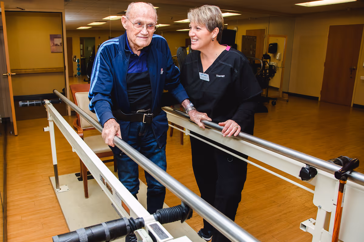 An elderly man wearing a blue jacket and jeans is walking with the support of parallel bars in a rehabilitation or therapy room. A female therapist in black scrubs stands beside him, smiling and providing encouragement. The room has wooden floors and beige walls with some chairs and therapy equipment in the background.
