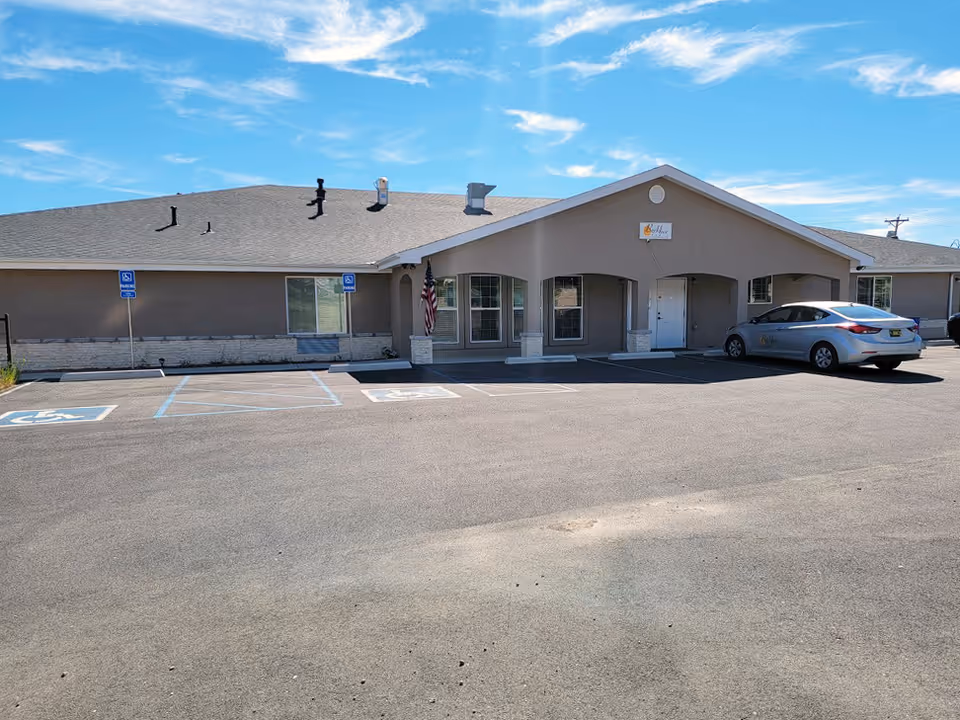 Front exterior of a single-story beige senior living building with a parking lot, handicap spaces, an American flag and a parked silver car.