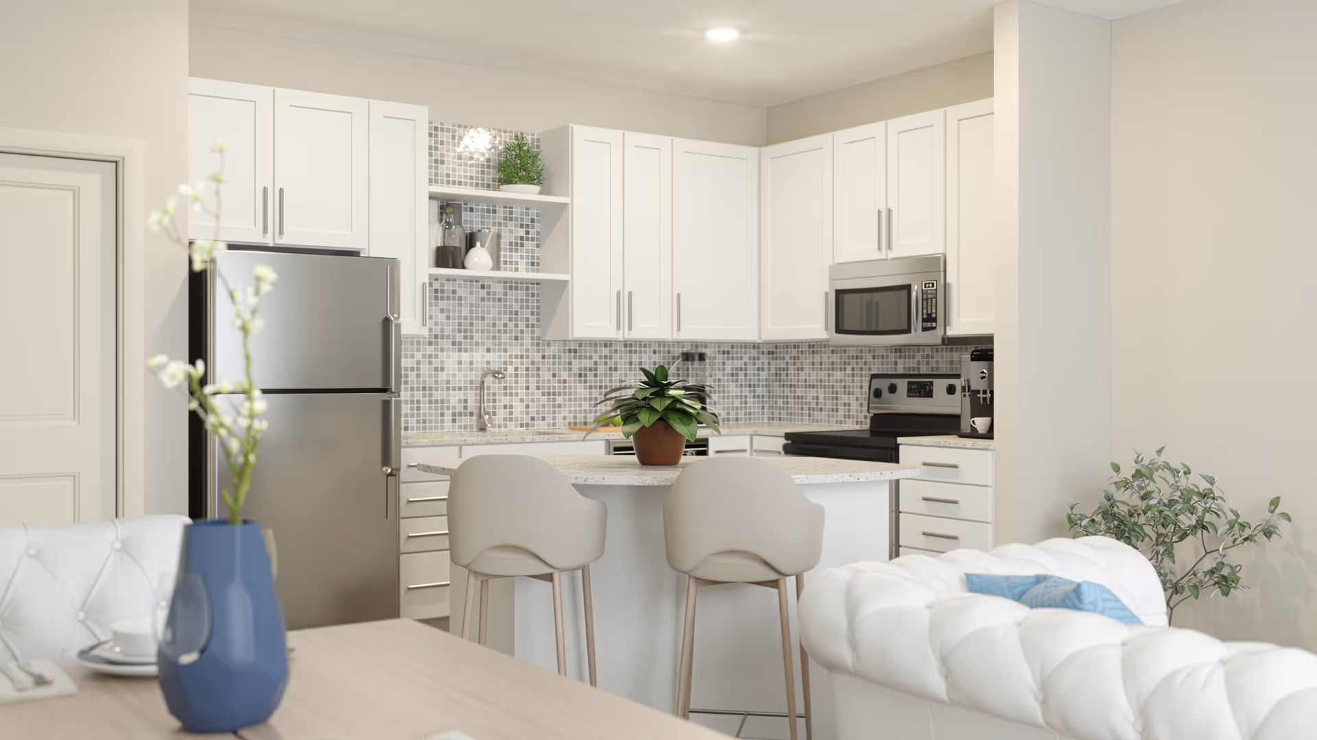 Modern kitchen with white cabinets, a mosaic tile backsplash, stainless steel refrigerator, microwave, and stove. There is a kitchen island with two beige bar stools and a potted plant on top. In the foreground, part of a white tufted sofa and a dining table with a blue vase holding white flowers are visible.