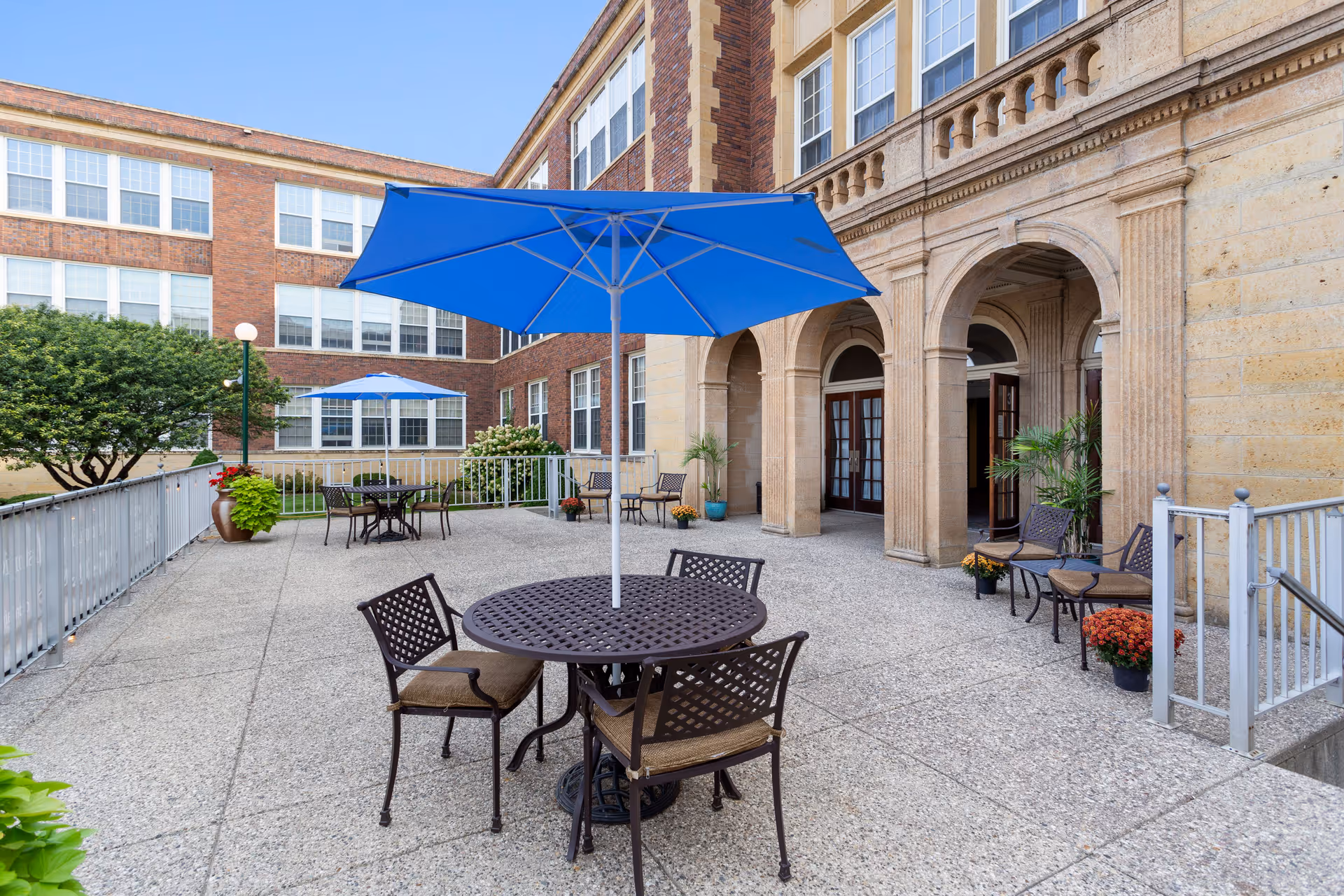 Outdoor patio area at Old Main Village with round metal tables and chairs, blue umbrellas providing shade, potted plants, and a building with large arched doorways and multiple windows in the background.