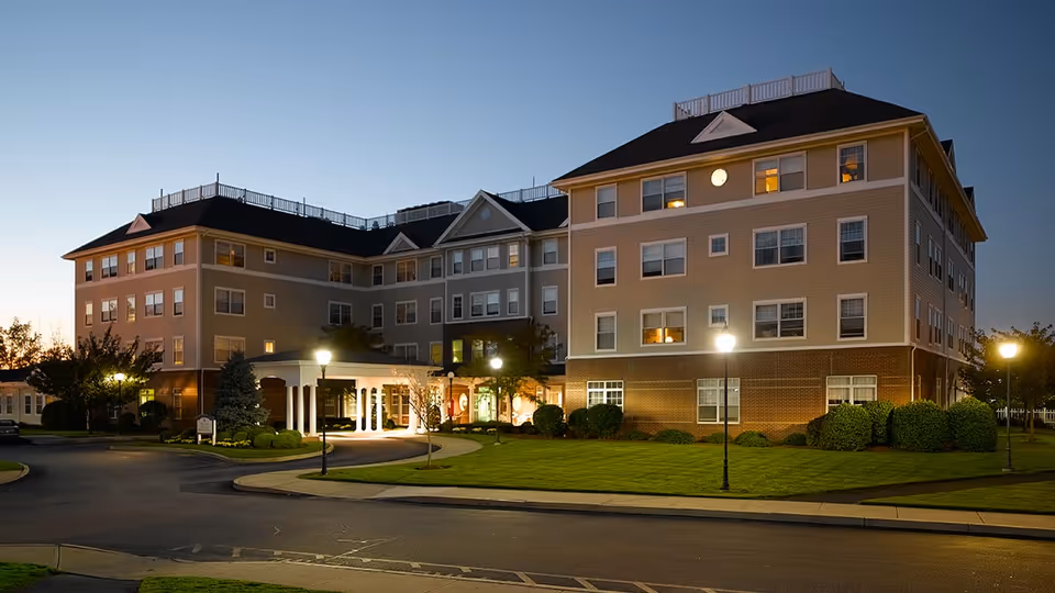 Exterior view of a multi-story senior living facility building at dusk with lights on inside and street lamps illuminating the entrance and surrounding lawn.