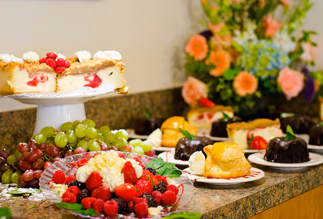 A dessert buffet with cakes, pastries, grapes and a platter of fresh berries arranged on a countertop with flowers in the background.