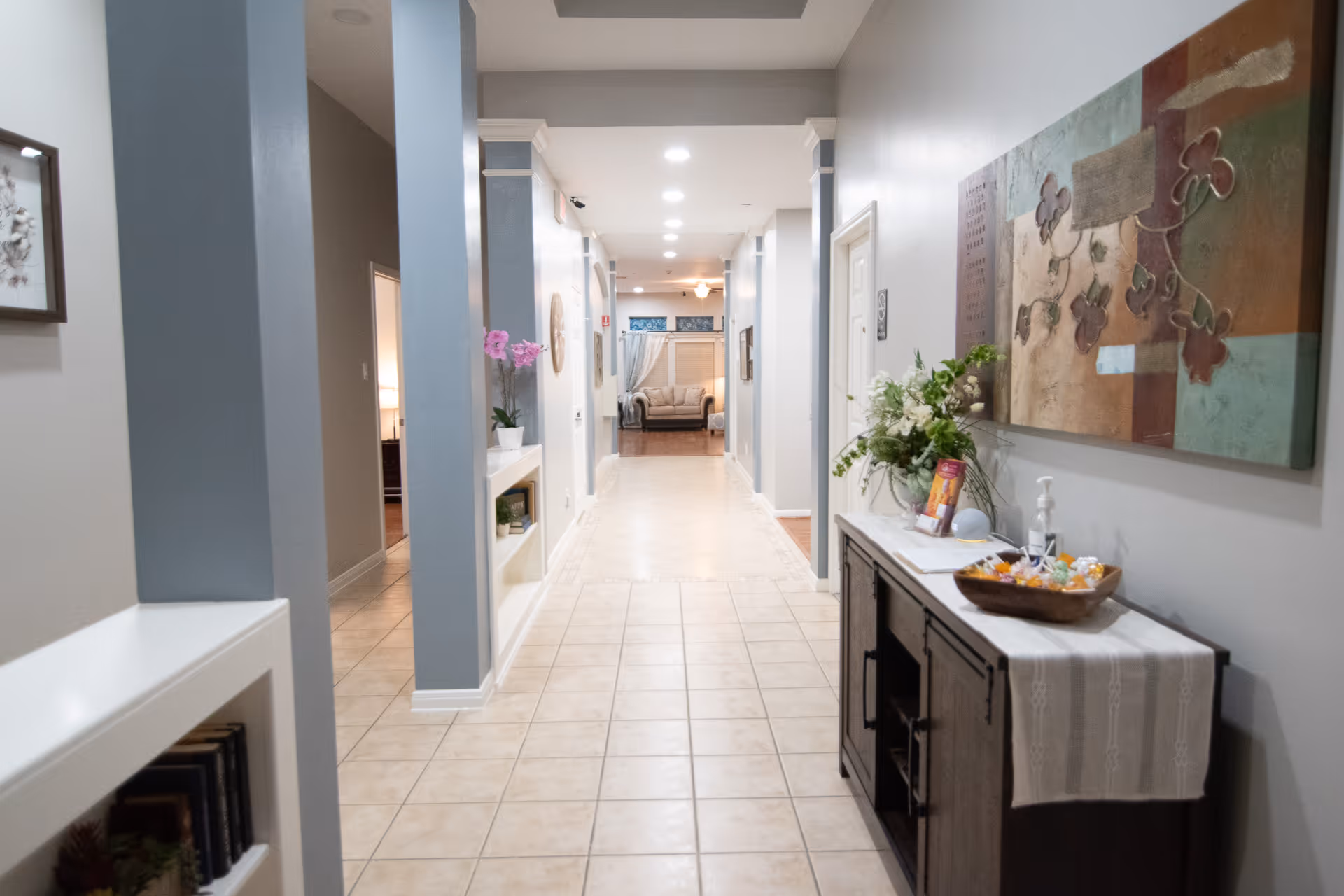 A well-lit hallway in a senior living facility with tiled floors and light-colored walls. On the right side, there is a dark wooden console table decorated with a floral arrangement, a bowl of candy, and a hand sanitizer dispenser. Above the table hangs a large abstract painting with floral motifs. On the left side, there are built-in shelves with books and a potted plant with pink flowers. At the end of the hallway, a cozy seating area with a sofa is visible.