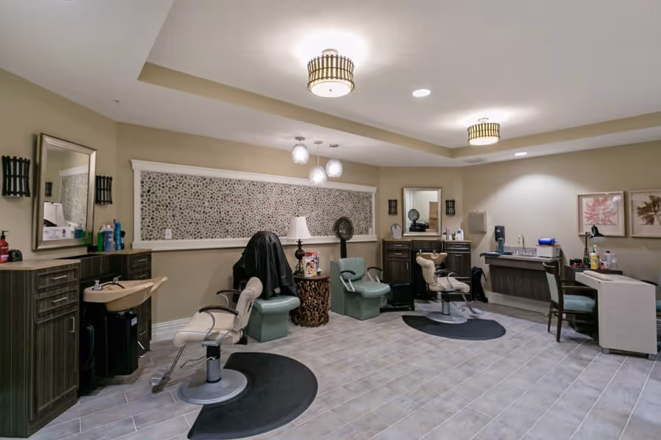 Interior of a salon area in an assisted living facility with multiple salon chairs, sinks for hair washing, mirrors, and a small table with a lamp. The room has beige walls, tiled flooring, and ceiling lights.